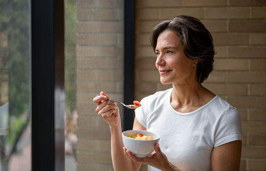 Happy woman at home eating a healthy breakfast while looking out the window - domestic life concept