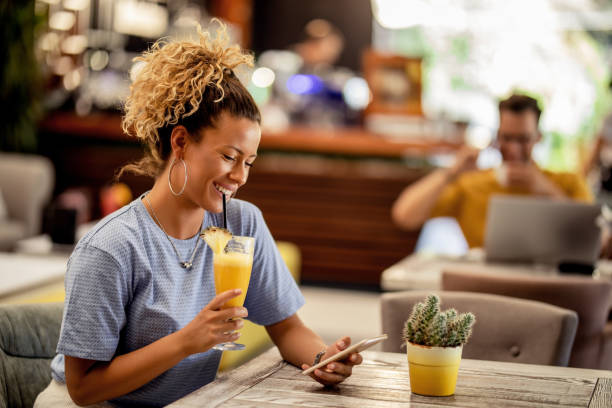 Happy woman drinking juice and using smart phone in a cafe. Young happy woman sitting in a bar and drinking fruit cocktail while text messaging on mobile phone. juice drink stock pictures, royalty-free photos & images