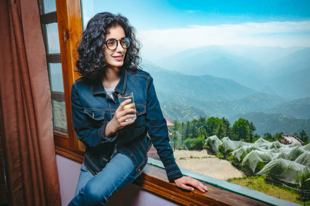 Happy woman enjoys scenic view through holiday villa's window. Indoor image of happy, Asian/Indian young woman, holds a glass of water, gives a toothy smile, sits on the window frame of a large glass window and looks at a beautiful scenic view of mountains at day time in a hill station, Shimla, Himachal Pradesh, India on her summer vacation. shimla stock pictures, royalty-free photos & images