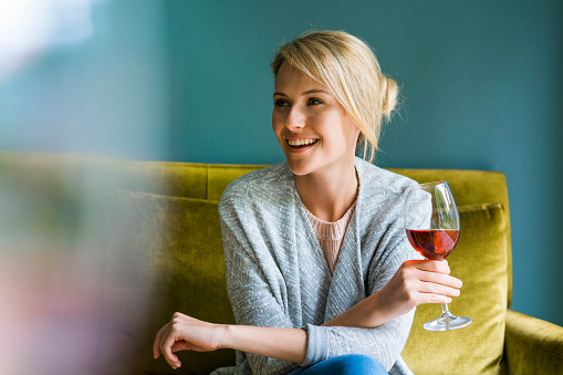 A photo of beautiful woman holding glass of red wine. Young female is looking away while sitting on sofa. Happy and relaxed lady is at home.