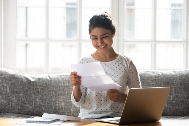 Happy woman holding paper reading good news college admission concept Indian ethnicity woman sitting on couch at home reading paper notice receive good news feels happy, cheerful student female looking at document enjoy exam results or college admission letter concept scholarships for females stock pictures, royalty-free photos & images