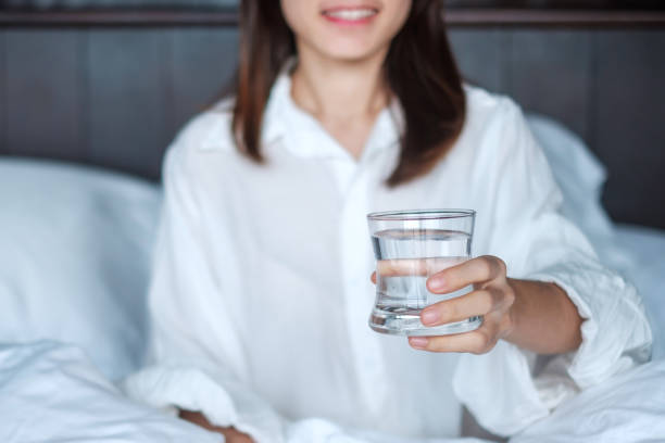 Happy woman holding water glass, female drinking pure water on bed at home. Healthy, Refreshment, lifestyle concept stock photo