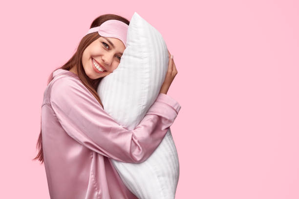 Happy woman hugging pillow in morning Side view of delighted young woman in nightwear smiling and embracing soft pillow after good night sleep against pink background eye mask stock pictures, royalty-free photos & images