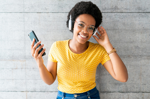 Portrait of a happy African American woman listening to music on her cell phone using headphones and looking at the camera smiling