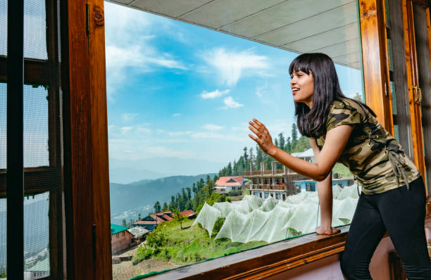 Happy woman looks at scenic view through holiday villa's window. Indoor image of happy, Asian/Indian young woman gives a toothy smile, looks through the large glass window with excitement and joy at a beautiful scenic view of mountains at day time in a hill station, Shimla, Himachal Pradesh, India on her summer vacation. shimla stock pictures, royalty-free photos & images