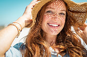 Portrait of beautiful woman wearing straw hat with large brim at beach and looking at camera. Closeup face of attractive smiling girl with freckles and red hair. Happy mature woman enjoing summer vacation at sea.