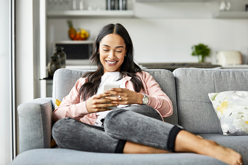 Smiling woman using mobile phone while sitting on sofa. Young female is text messaging in living room. She is relaxing at home.