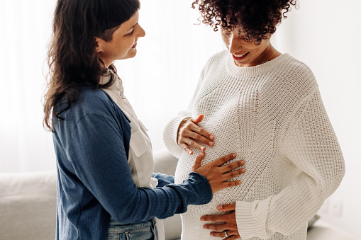 Happy young woman touching a surrogate mother's belly bump. Woman smiling while feeling the movement of a pregnant woman's baby. Young woman spending time with her surrogate at home.