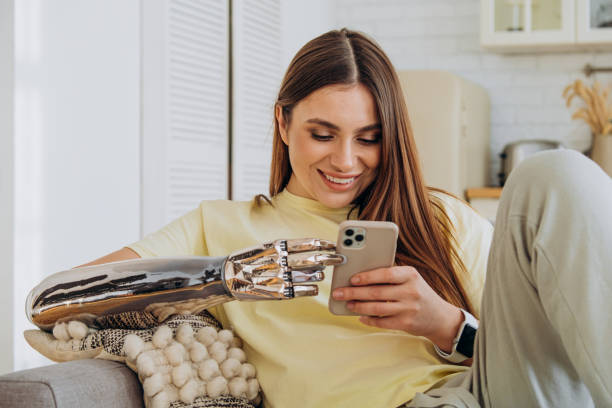 A happy woman with a bionic prosthetic arm sits on the couch with a phone and communicates online with friends or surfs the Internet or makes online purchases stock photo