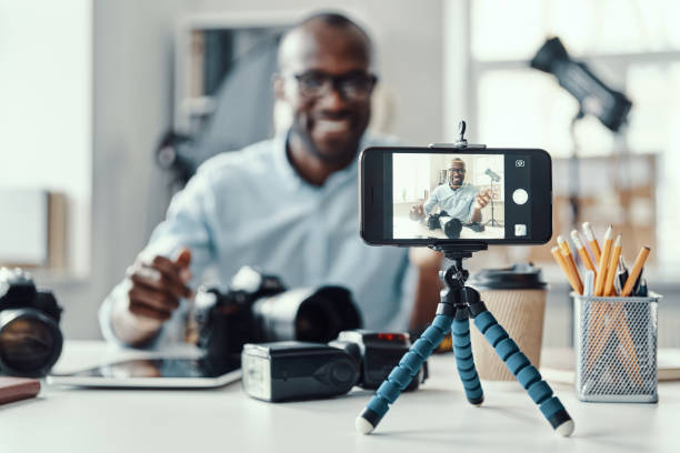 Happy young African man in shirt Happy young African man in shirt showing digital camera and telling something while making social media video vlogging stock pictures, royalty-free photos & images