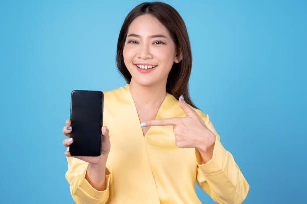 Happy young Asian beautiful woman holds a cell phone and a finger pointing at the phone screen, Isolated over blue background. stock photo