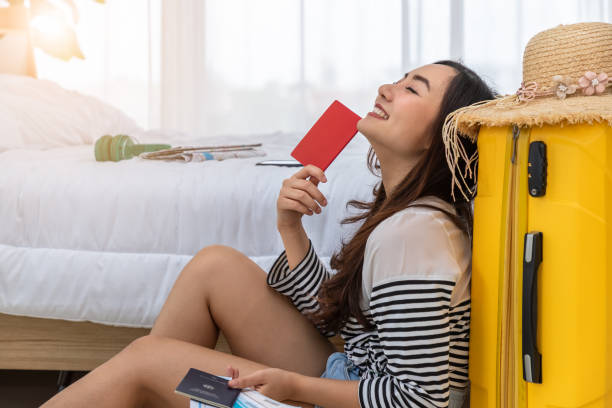 Happy young Asian woman sitting with luggage, preparing suitcase for summer vacation travel trip, holding passport and ticket, closed eyes, dreaming. stock photo