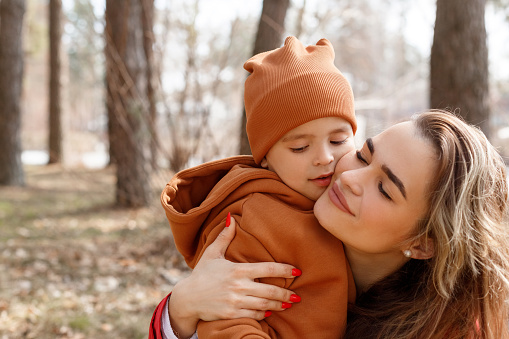 Happy young beautiful mom with closed eyes hugs her son tenderly and heartwarming. Sunny family portrait in the forest, spring or autumn
