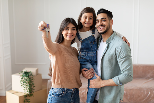 Portrait Of Happy Young Family Showing Key, Cheerful Parents Hugging Little Kid, Holding Daughter On Hands. Smiling People Moving In New Apartment Standing In Living Room. Selective Focus