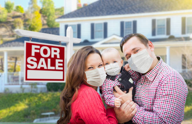 Happy Young Family Wearing Medical Face Masks In Front of New House and Sold For Sale Sign stock photo