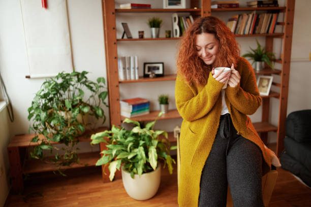 A happy young ginger girl is standing in the living room in the morning and enjoying her morning coffee. stock photo