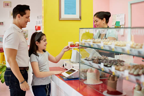 Happy Young Girl Taking Plate of Cupcakes from Bakery Worker Side view of Hispanic father in late 40s and 9 year old daughter smiling as woman hands them a plate of fresh gourmet cupcakes in Miami bakery. small business saturday stock pictures, royalty-free photos & images