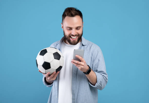 Happy young guy with soccer ball using smartphone, winning sports bet, rooting for his favorite team on blue background Happy young guy with soccer ball using smartphone, winning sports bet, rooting for his favorite team on blue studio background. Millennial man watching online football championship sports betting stock pictures, royalty-free photos & images