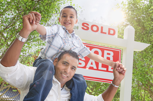 Happy Young Hispanic Father and Son In Front of Sold For Sale Real Estate Sign.