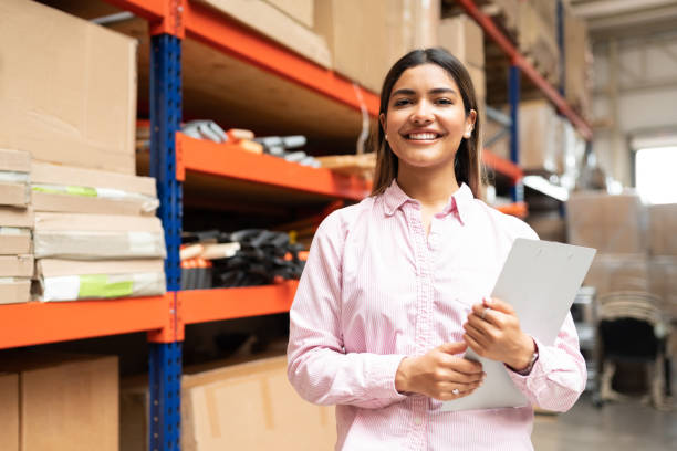 Happy Young Latin Worker With List Working At Factory Portrait of pretty smiling female employee holding inventory clipboard while standing against rack at warehouse mexican woman stock pictures, royalty-free photos & images