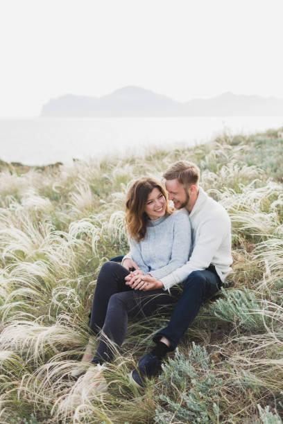 Happy young loving couple sitting in feather grass meadow, laughing and hugging, casual style sweater and jeans stock photo