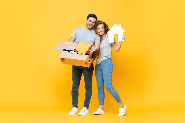 Happy young man carrying stuff standing with his girlfriends in yellow isolated studio background, moving house concept stock photo