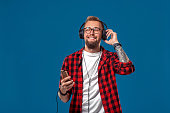 Happy young man listening to music with headphones. Handsome smiling guy in checkered shirt with closed eyes dancing with headphones. Isolated on blue background. Concept of people emotions