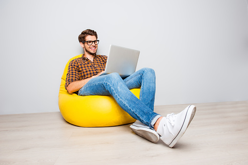 Happy young man sitting in yellow pouf  and using laptop