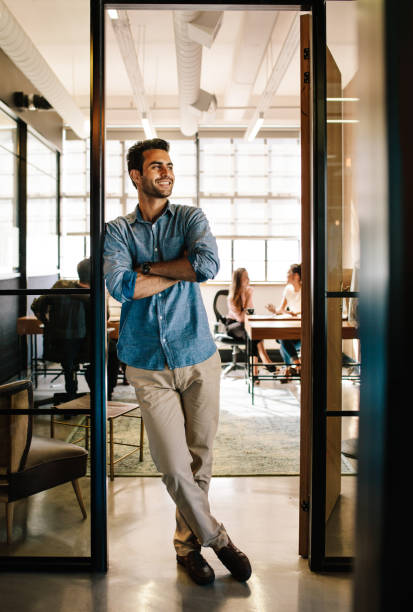 Happy young man standing in office doorway Full length portrait of happy young man standing in doorway of office with his arms crossed and looking away. Male executive at office with people working in background. leaning stock pictures, royalty-free photos & images