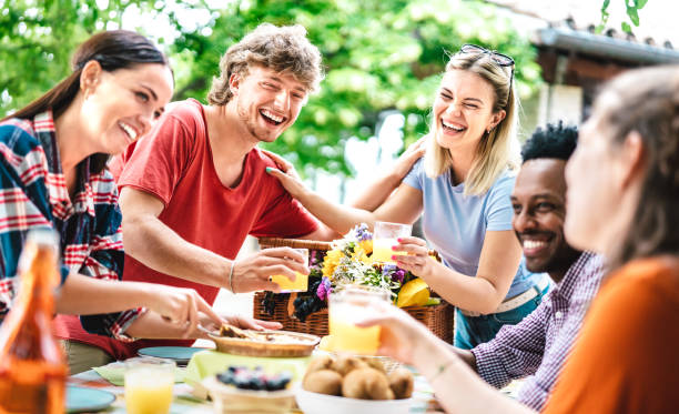 Happy young men and women toasting healthy orange fruit juice at farm house picnic - Life style concept with alternative friends having fun together on afternoon relax time - Bright vivid filter stock photo