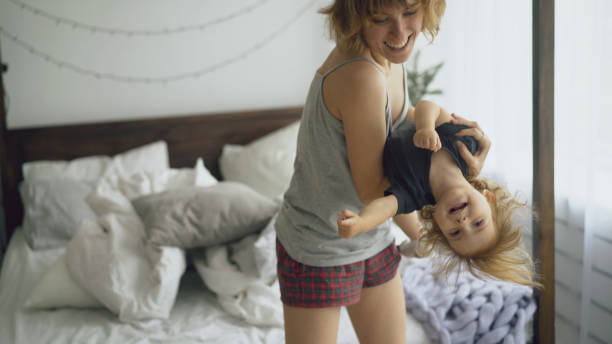 Happy young mother and little cute daughter dancing near bed in bedroom at home stock photo