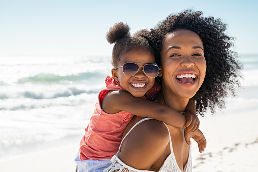 Smiling black mother and beautiful daughter wearing sunglasses having fun on the beach. Portrait of happy african american woman giving a piggyback ride to her cute little girl wearing shades while looking at camera. Kid embracing her bigger sister during summer vacation with copy space.