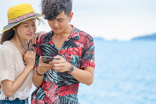 Happy young smart Asian couple man and woman standing on rocky seacoast beach together using smartphone gadget with internet wireless technology for social media or online shopping in summer vacation