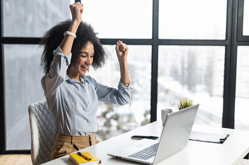 African-American woman with curly hairstyle hands up,at the desk with open laptop,wide smile,excited by the news,celebrating her success,accepted for internship,admitted to university, got a letter