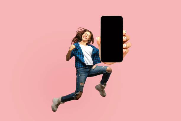 Happy young woman flying and jumping in air and showing big mobile empty screen stock photo
