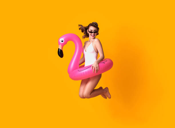 glückliche junge frau springt auf gelbem hintergrund in bademode mit flamingo gummiring strand gekleidet. - badebekleidung stock-fotos und bilder