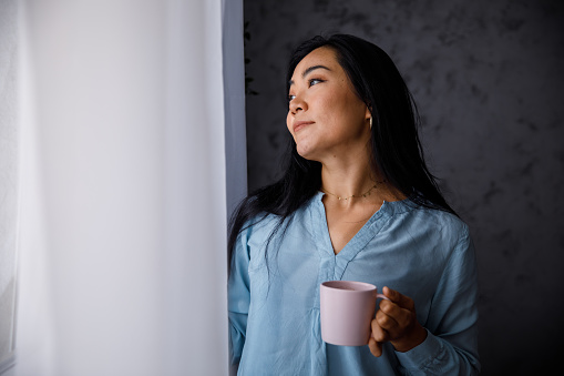Portrait of beautiful, young Mongolian woman standing by the window, holding a cup of coffee she is enjoying, looking outside and contemplating.