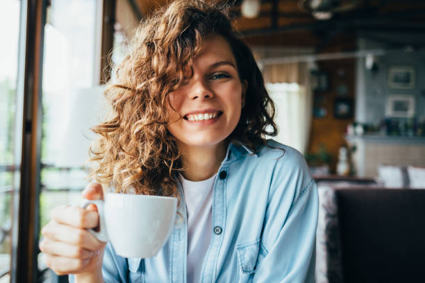 Happy young woman with curly hair Happy young woman with curly hair smiling holding white cup of coffee sitting at table by the window in restaurant in daytime. curley cup stock pictures, royalty-free photos & images