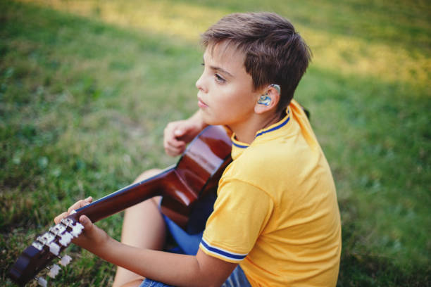 niedosłyszący chłopiec grający na gitarze na świeżym powietrzu. dziecko z aparatami słuchowymi w uszach gra muzykę i śpiewa piosenkę w parku. hobby artystyczne dla dzieci. autentyczny moment dzieciństwa. - hearing aid zdjęcia i obrazy z banku zdjęć