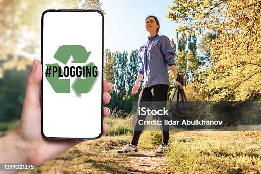 istock Hashtag plogging. Close up of female hand showing smartphone. Background with a woman at the park holding a bag of garbage. The concept of volunteering and environmental protection 1399321275