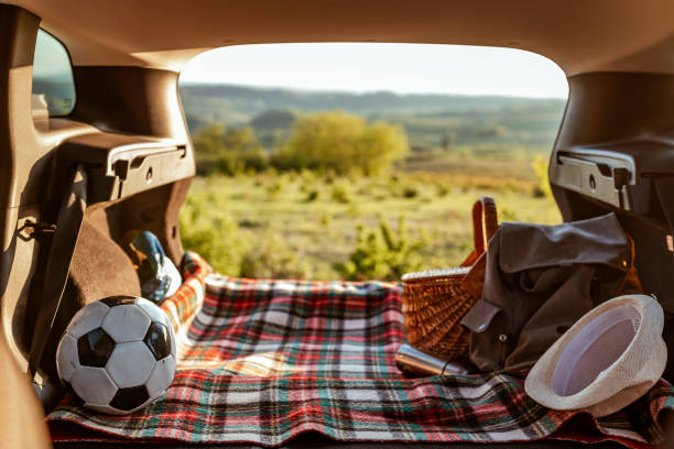 Photo of a family car ready for the summer holiday. The trunk of SUV packed for a weekend road trip. Photo of interior of open car trunk and garden. Soccer ball and picnic basket in a car trunk on a sunny day.