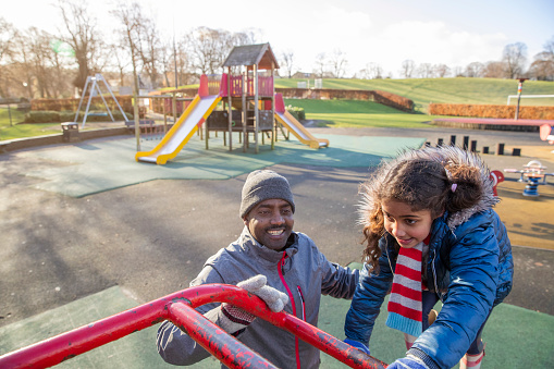 A mature black man and his mixed race daughter wearing warm casual clothing. He is helping her while she climbs up a jungle gym in a play park on a sunny winters day.