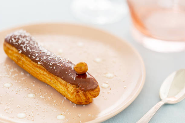 Hazelnut and chocolate French eclair pastry on pink plate, aside a pink vase, a spoon and a glass over a light pastel green napkin. stock photo