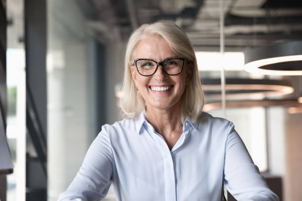 Headshot of middle-aged businesswoman posing at workplace Headshot of smiling middle-aged businesswoman in glasses look at camera pose shooting live webinar broadcast in office, happy mature female employee talk on video call at workplace, leadership concept businesswoman stock pictures, royalty-free photos & images