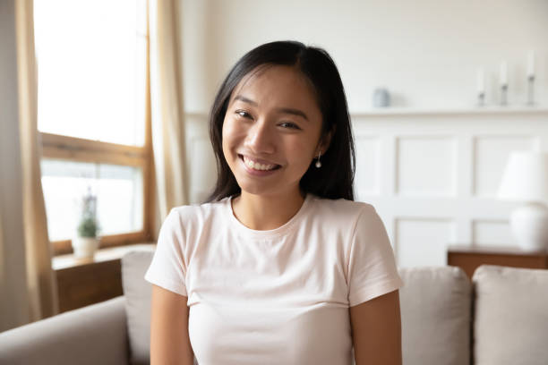 Headshot of millennial Asian woman have video call Headshot portrait of smiling Asian millennial girl sit on couch look at screen having video call at home, happy Vietnamese young woman talk speak using online dating service on gadget or device injecting photos stock pictures, royalty-free photos & images