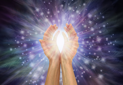 female hands with a white healing energy burst between against radiating sparkles and feathers dark blue background with copy space