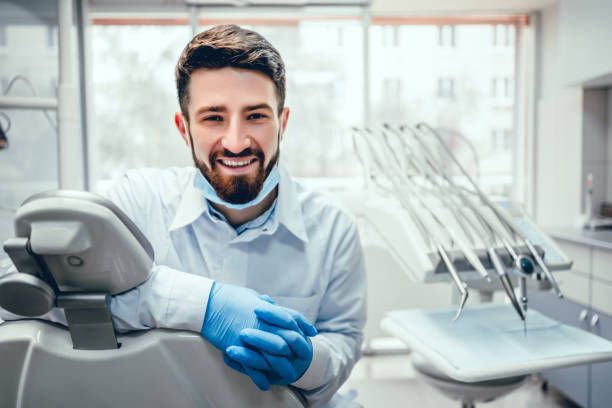 Healthcare and medicine concept. Front view of professional male dentist in white doctor coat and protective gloves sitting in dental chair and equipment, looking at camera and smiling. Bearded man posing during working process. dentist stock pictures, royalty-free photos & images