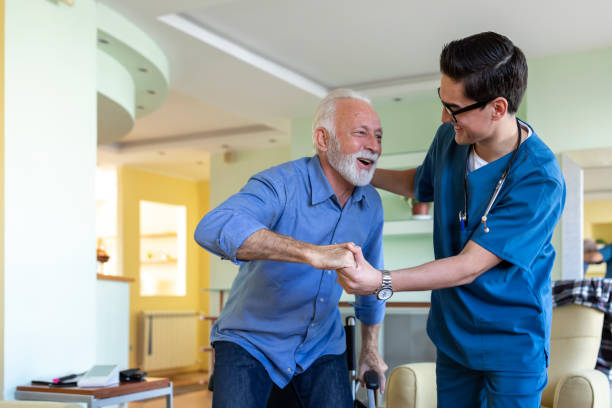Healthcare Worker is in Home Visit to an Older Man with Physical Disabilities and Assisting him to Get Up. Senior Man in a Wheelchair is Having a Positive Communication with a Young Doctor in Blue Uniform who is Giving him Support and Helping Him to Get Up From a Wheelchair. physician assistant stock pictures, royalty-free photos & images