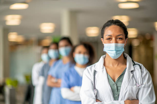 Healthcare workers portrait A diverse group of women stand in an institutional building in medical wear. A woman wearing scrubs stands at the forefront with a mask that is pulled down. female doctor stock pictures, royalty-free photos & images