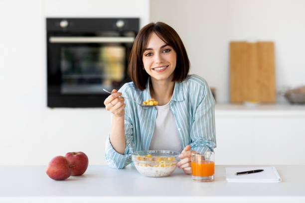 concepto de desayuno saludable. feliz dama comiendo muesli bowl con manzanas y jugo, sentada en el interior de la cocina moderna - avena fotografías e imágenes de stock